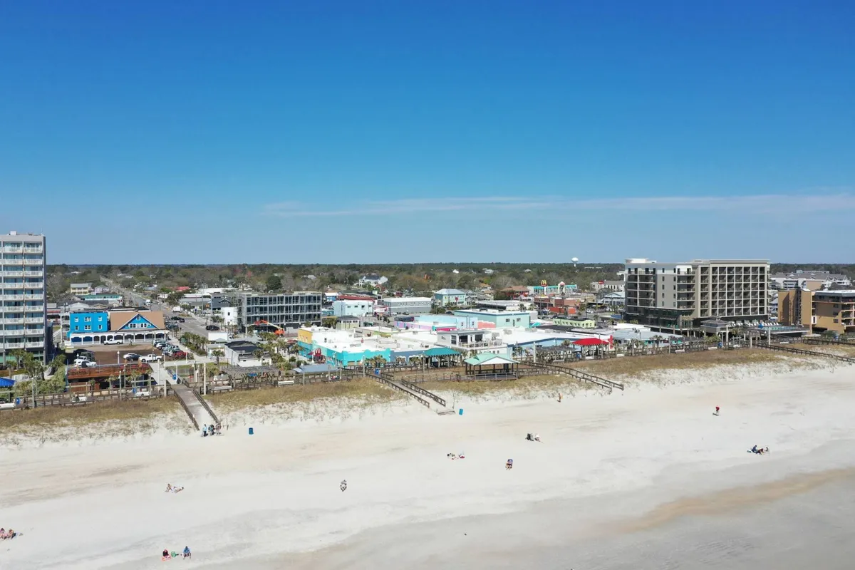 A wide shot of the beach featuring colorful buildings, the boardwalk, and groups of people relaxing or engaging in beach activities. The image conveys a joyful, social vibe with shopping, dining, and sunset events.