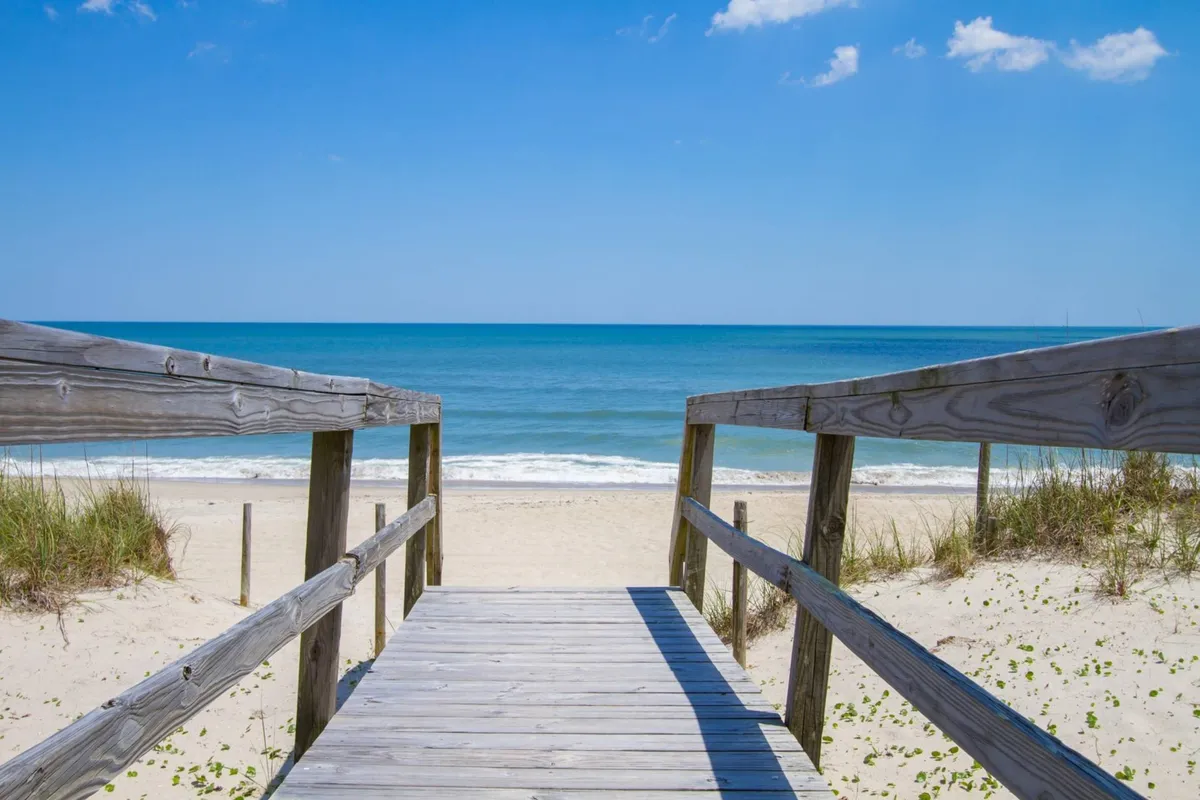 A wide wooden walkway leads directly to the inviting sandy beach, with the blue ocean stretching out under a sunny sky. The scene features gentle waves, beachgoers enjoying the sand and surf, and a sense of space for family activities like building sandcastles.