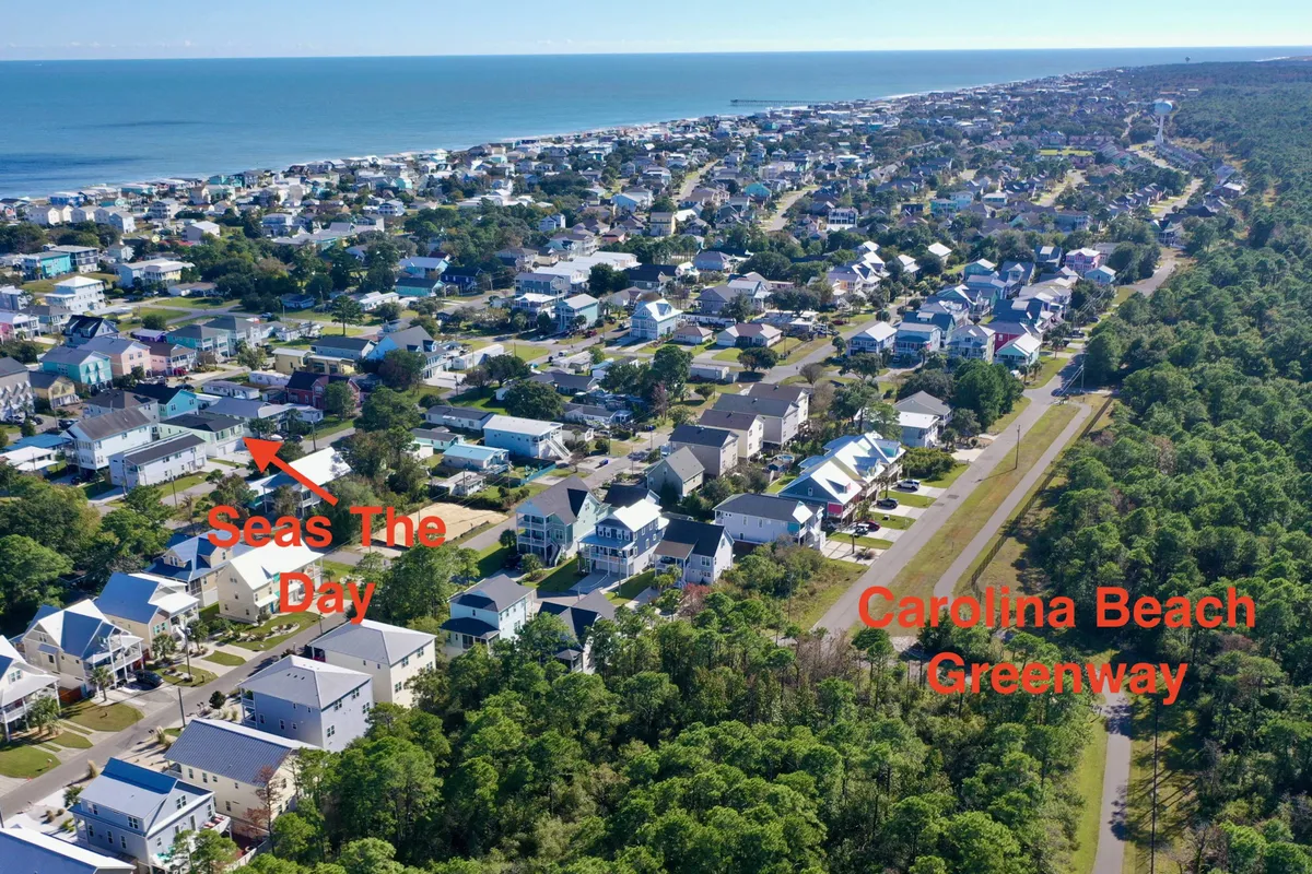 A view of residential homes lining a greenway path that runs alongside the beachfront, with the ocean shimmering in the background. The greenway invites biking and walking, while shady trees offer spots for picnics and relaxation.
