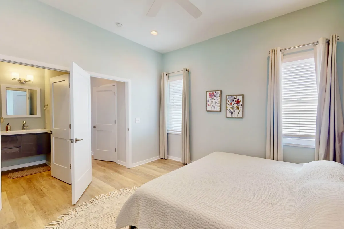 A tranquil bedroom featuring a plush bed with fresh linens, soft accent pillows, and a peaceful color scheme. The room includes a doorway leading to a private ensuite bathroom and tasteful wall art, with natural light streaming through a window.
