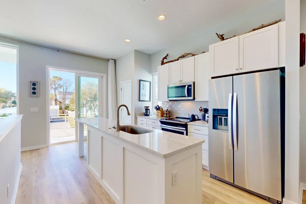 A modern kitchen with a prominent kitchen island, stainless steel appliances, and large windows that fill the space with welcoming natural light. The open doorway hints at easy indoor-outdoor flow, and the space is styled with sleek, contemporary décor.