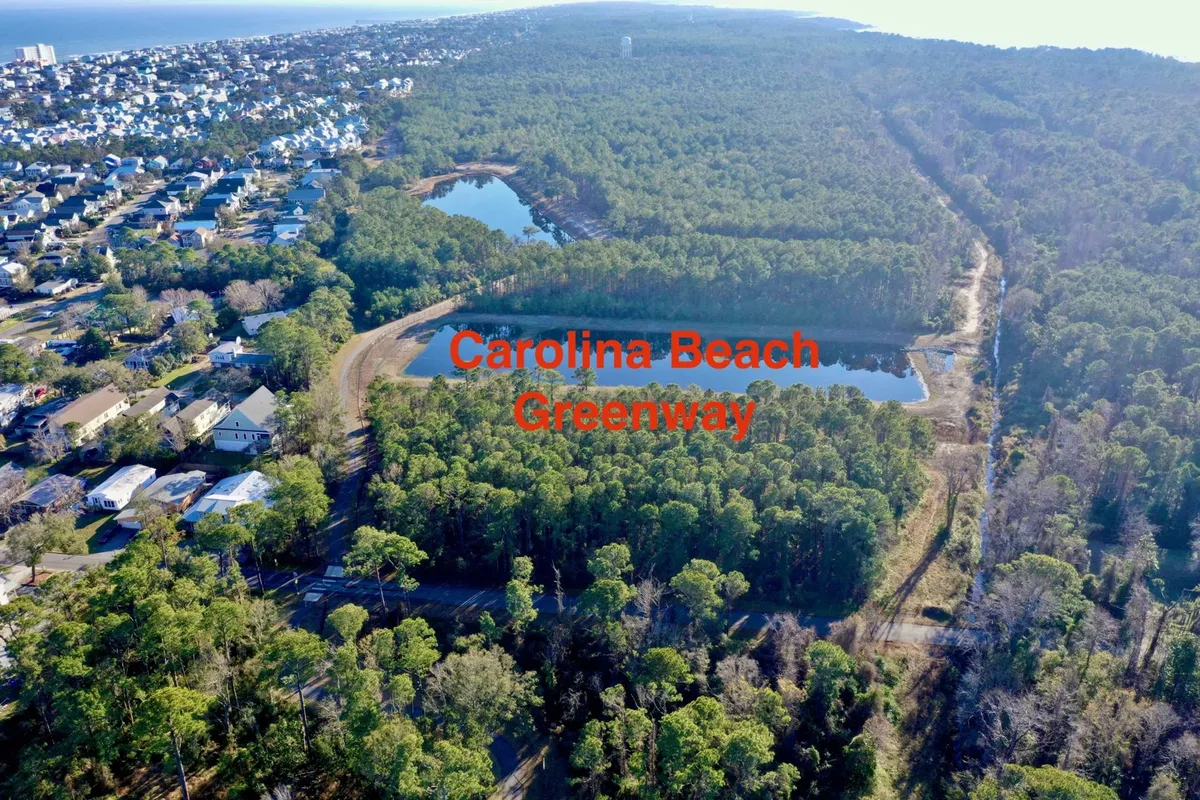 The image presents a sweeping view of lush greenery interspersed with bodies of water, neighboring residential areas, and a wide open coastal vista. The landscape is ideal for outdoor activities such as hiking and biking, with tranquil ponds and blooming wildflowers adding to the natural beauty.