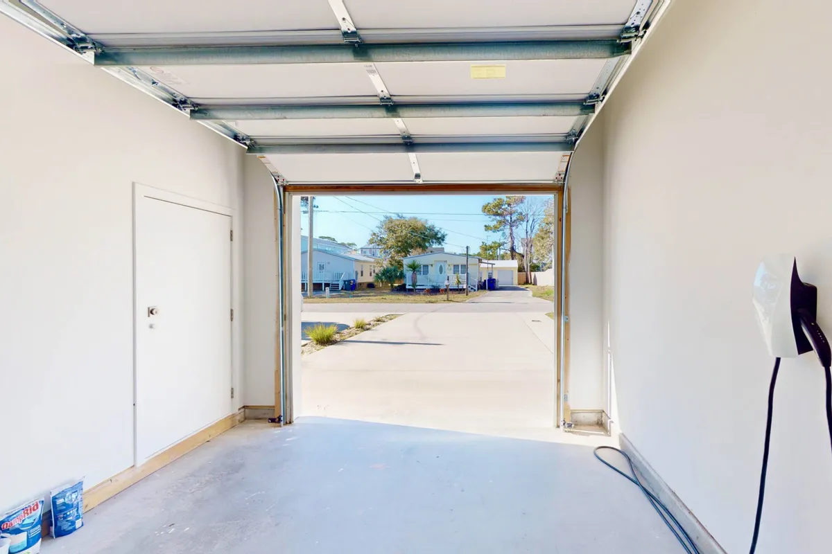 A spacious, clean garage with an open garage door, concrete floor, and side door visible. The area appears uncluttered, offering plenty of room for storing vehicles, adventure gear like bikes or kayaks, and other outdoor equipment. The openness and light suggest a practical, functional space ideal for families or groups preparing for beach outings or excursions.