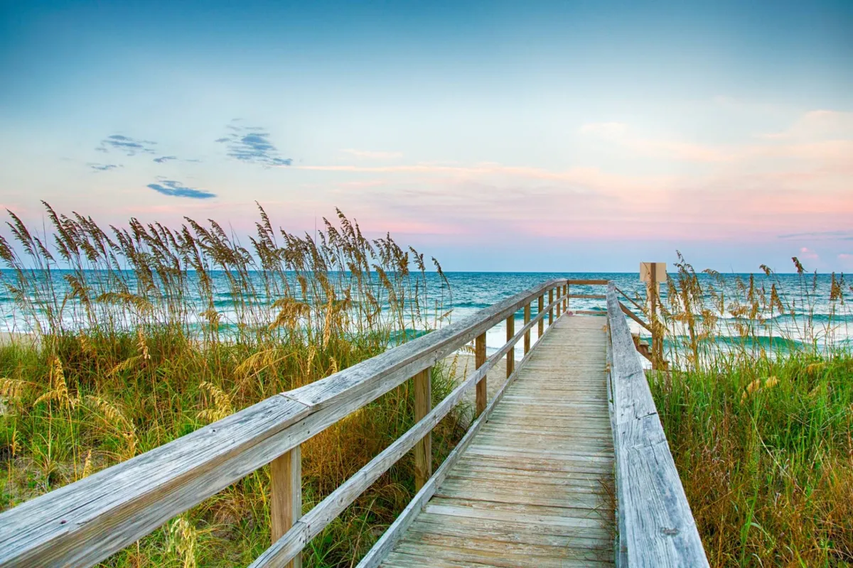 A wooden boardwalk stretches over dune grass toward the ocean, under a colorful sunset sky. The scene is tranquil and open, suggesting opportunities for beach walks, photography, and enjoying the coastal atmosphere.