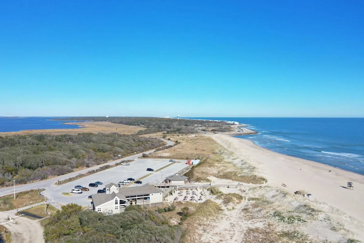 A tranquil exterior view of the beach and ocean with a clearly visible parking area, a nearby restaurant, and surrounding natural scenery. The setting feels inviting for those arriving by car, with ample space and a relaxed coastal vibe.