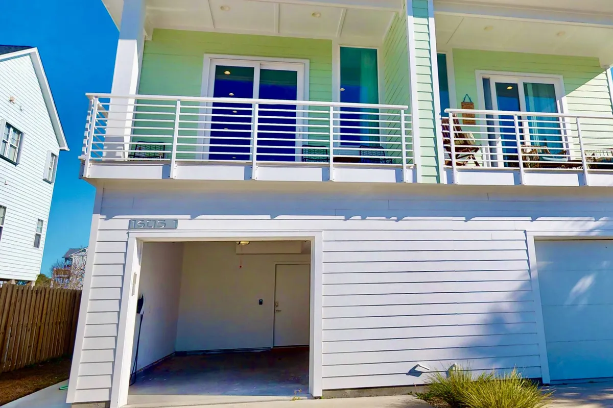 An exterior shot showing the green-sided building with a visible balcony and the entrance to the garage at ground level. The driveway in front offers additional parking and glimpses of the property’s curb appeal, making it clear there is space for multiple vehicles and easy access for coming and going.