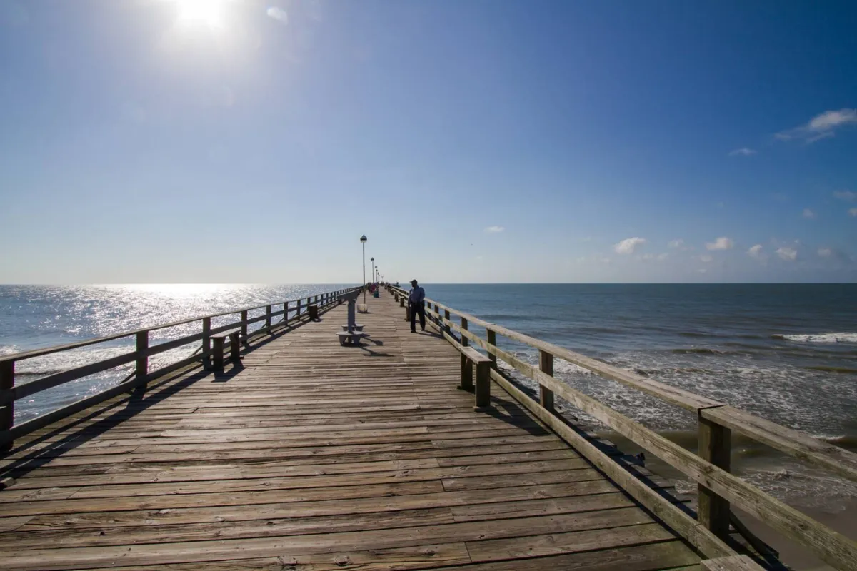 A scenic exterior view of the wooden pier at Kure Beach, stretching into the ocean under sunlight with people strolling along it. The atmosphere is peaceful yet active, perfect for group outings and daytime recreation.